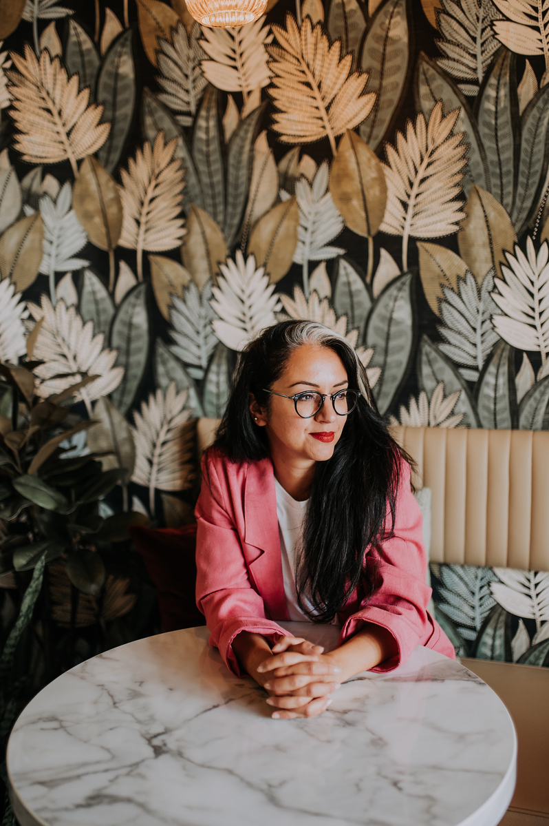 A woman in a restaurant-dining room-feature foliage wallpaper-marble topped table-cream leather banquet seating-pink blazer